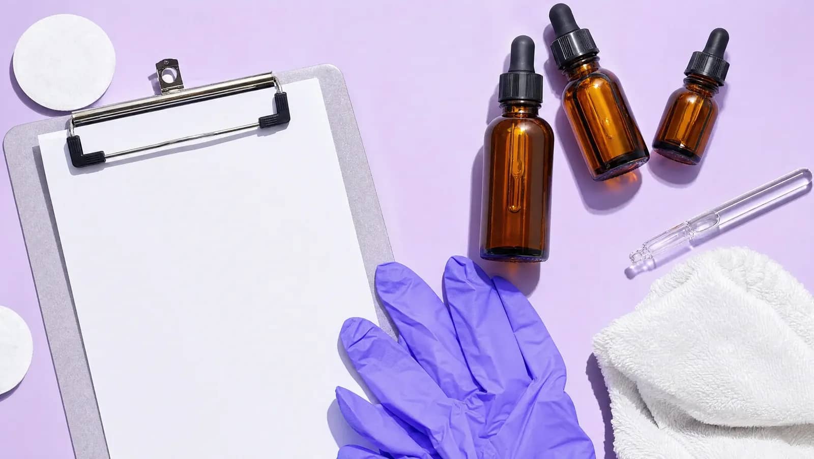 Prancheta em branco, luvas dobradas, algodão e frascos âmbar com pipeta vistos de cima sobre fundo lavanda