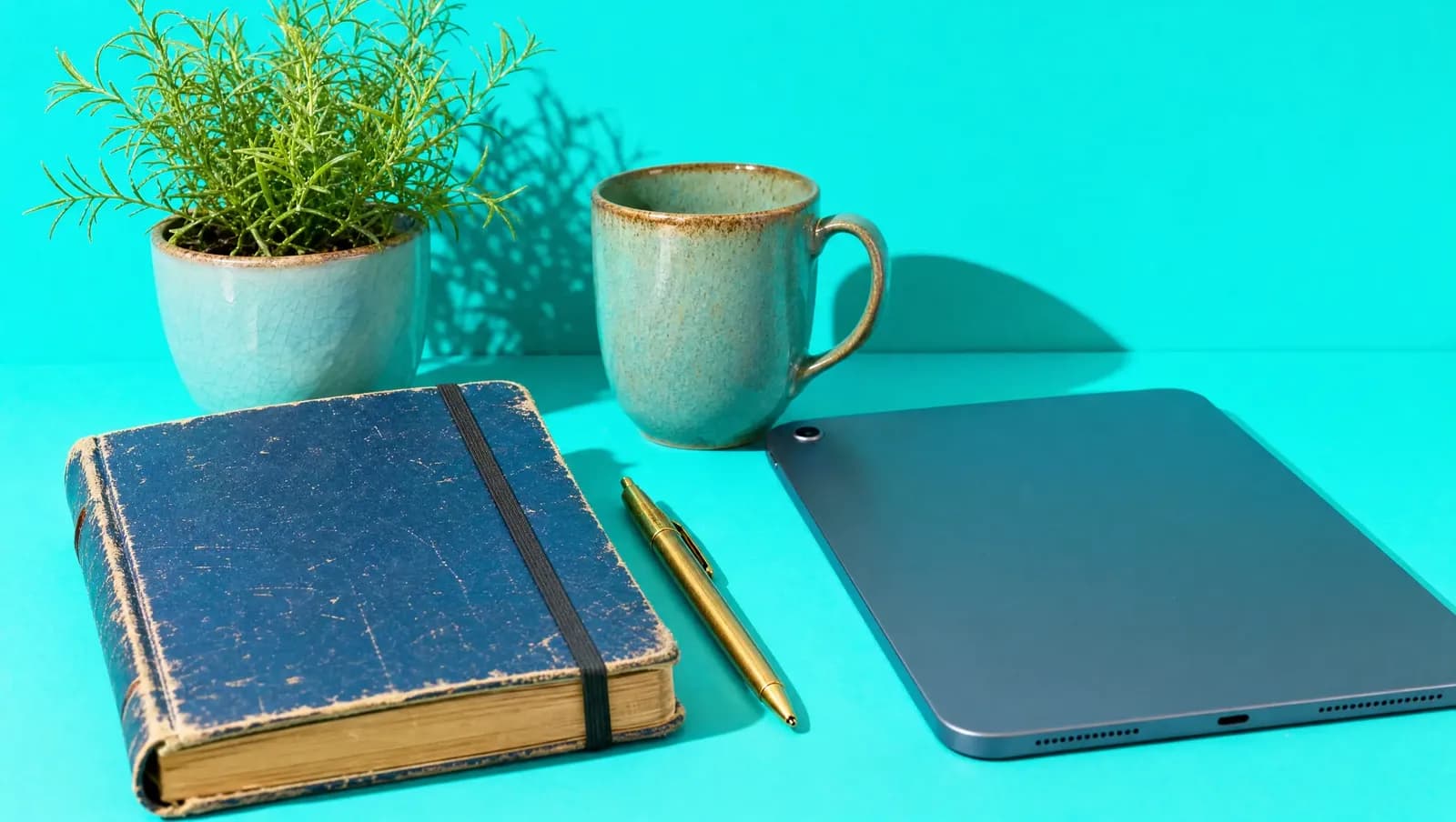 Caderno velho fechado com elástico ao lado de um tablet virado para baixo, com caneta e café, sobre fundo turquesa