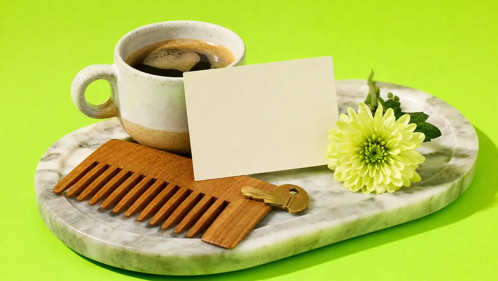 Xícara de café, cartão em branco, pente de madeira, chave de latão e uma flor em bandeja de mármore, fundo verde