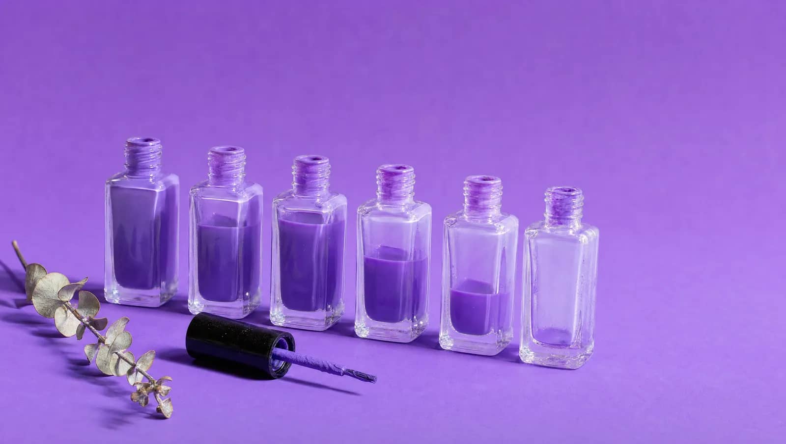 Seis vidros de esmalte idênticos enfileirados sobre fundo violeta, o primeiro cheio e cada seguinte com menos esmalte lavanda, até o sexto vazio; à frente, a tampa com o pincel deitada, e à esquerda um ramo de eucalipto seco