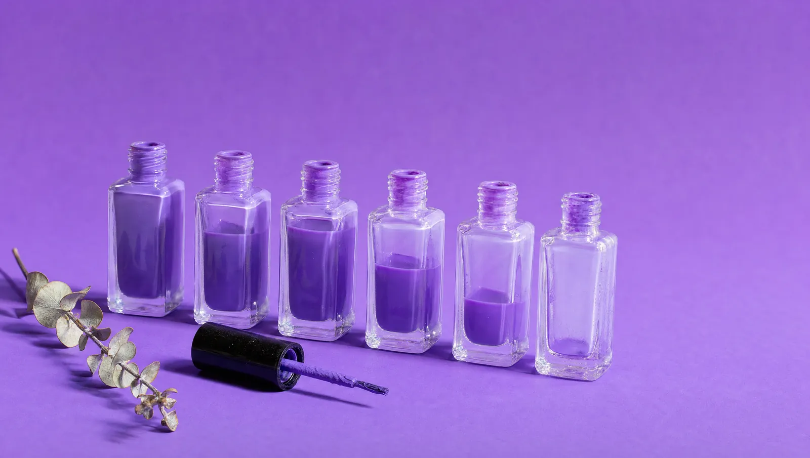 Seis vidros de esmalte idênticos enfileirados sobre fundo violeta, o primeiro cheio e cada seguinte com menos esmalte lavanda, até o sexto vazio; à frente, a tampa com o pincel deitada, e à esquerda um ramo de eucalipto seco