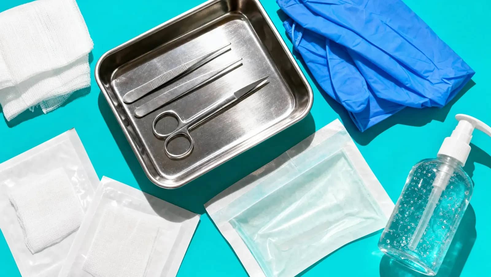 Bandeja de inox com pinças e tesouras, luvas, gaze estéril e álcool em gel vistos de cima sobre fundo turquesa
