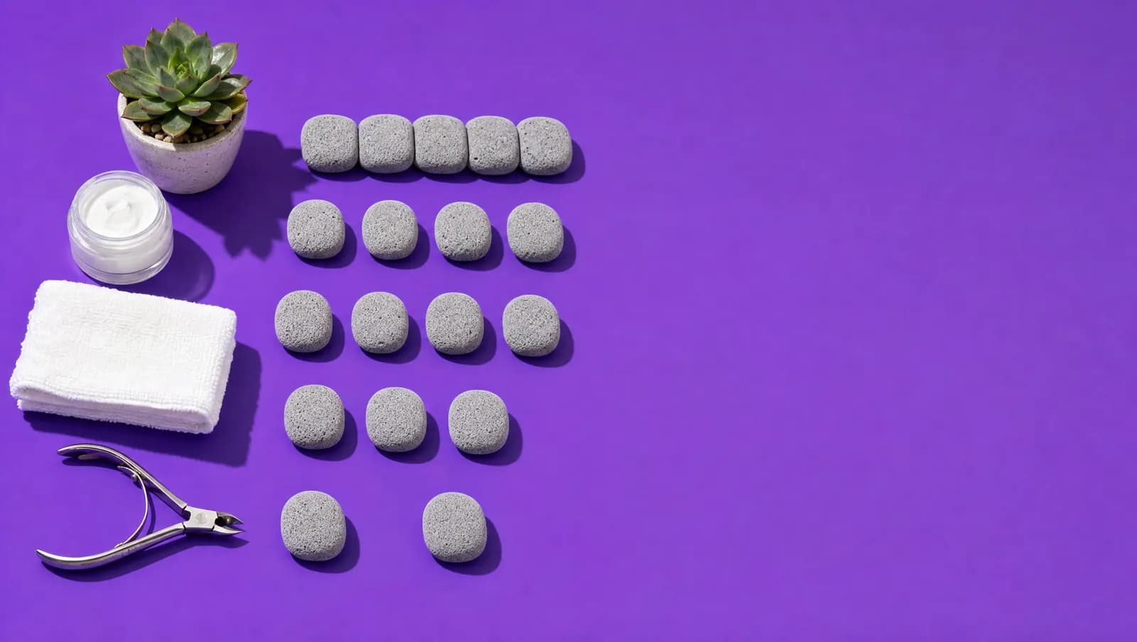 Fotografia de cinco fileiras de pedras-pomes sobre fundo violeta, com os espaços entre as pedras aumentando de uma fileira para a outra, ao lado de um alicate de podologia, um pote de creme e uma suculenta