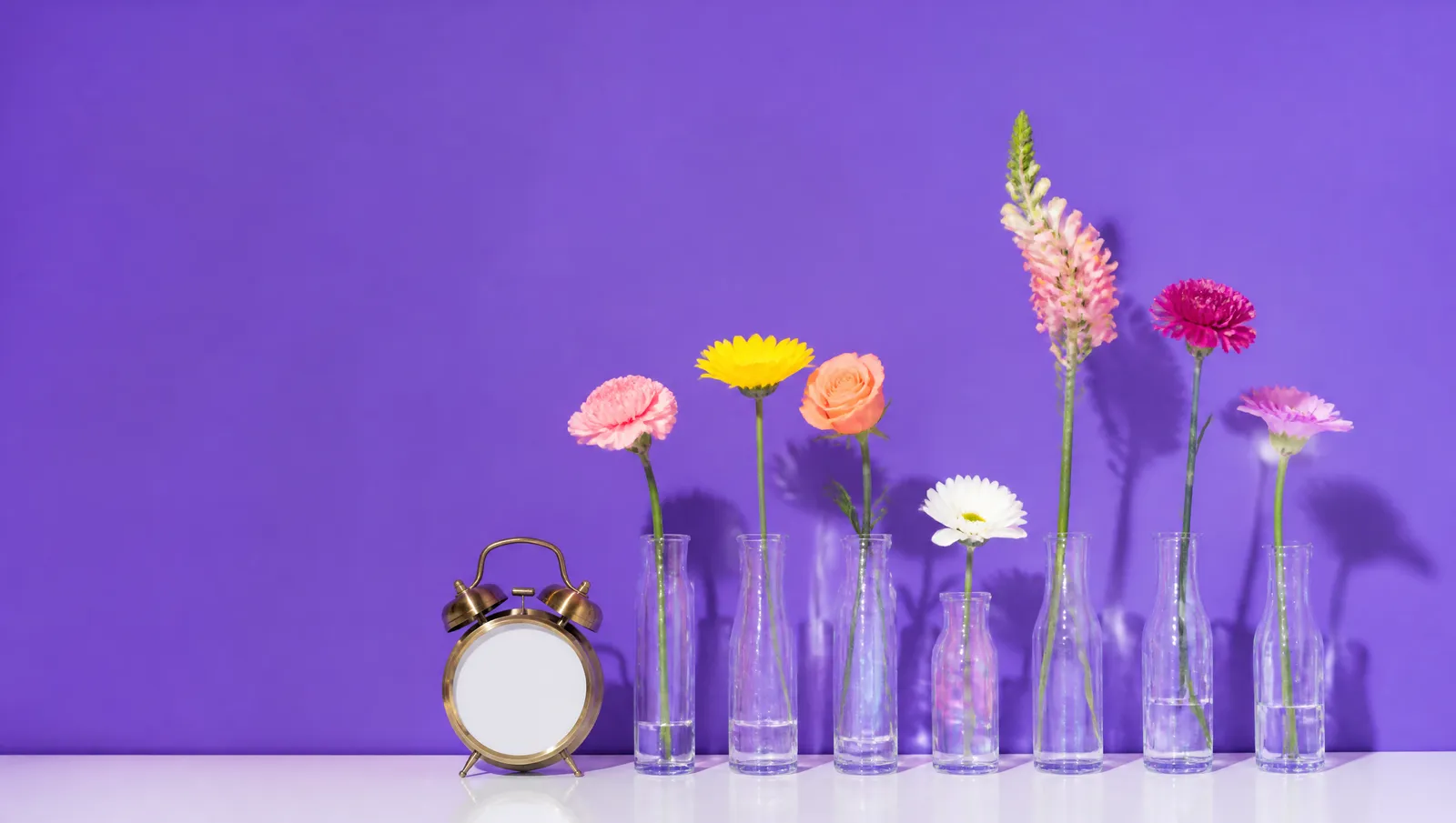 Sete vasos de vidro com flores de alturas diferentes desenhando a curva de um dia — sobe, cai no meio, atinge o pico e desce — ao lado de um despertador dourado de face lisa, sobre fundo violeta