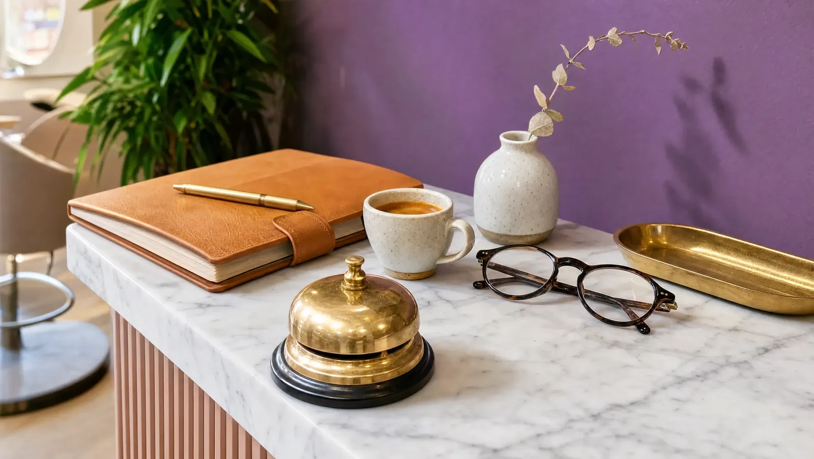 Balcão de recepção em mármore com campainha de latão, agenda de couro, café e óculos, diante de parede violeta
